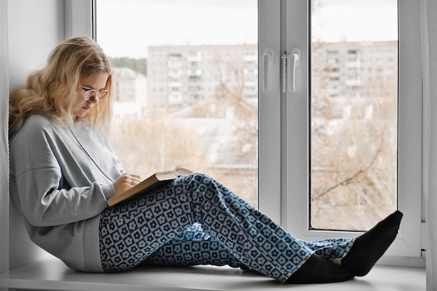 Beautiful girl in casual clothes sits on windowsill and reading book