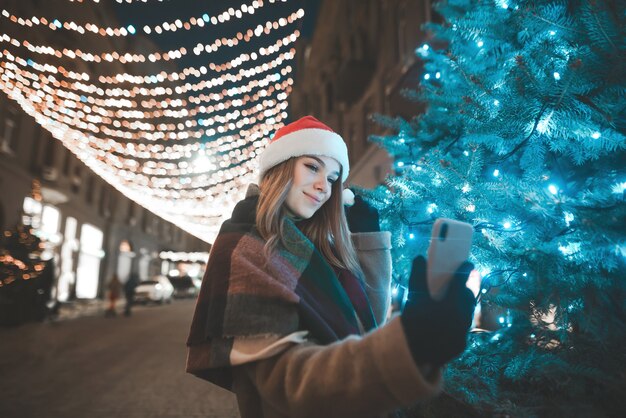 Beautiful girl in a Christmas hat stands at night on a street near a Christmas tree and takes a selfie