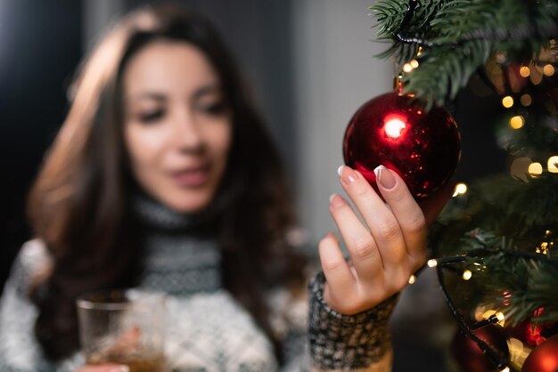 Beautiful girl in a Christmas sweater stands near the Christmas tree with a glass of champagne in her hand Touches a toy on the Christmas tree Focus on the ball Celebration of new year christmas