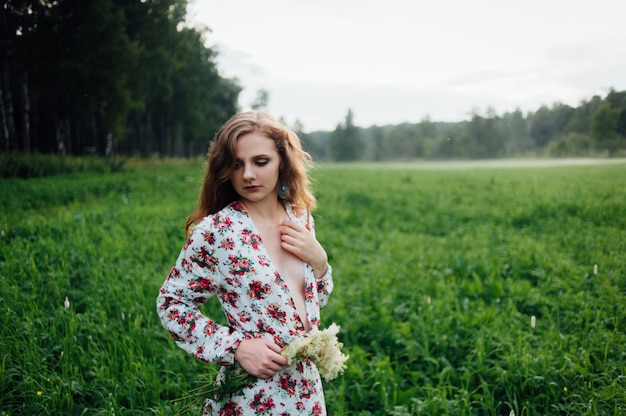 A beautiful girl in a colorful dress stands in the evening on a meadow against a background of fog. 