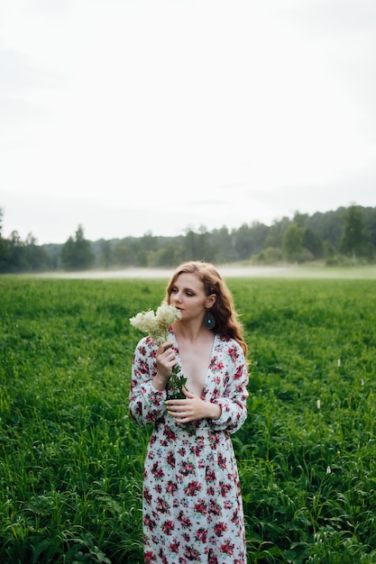 A beautiful girl in a colorful dress stands in the evening on a meadow against a background of fog. 