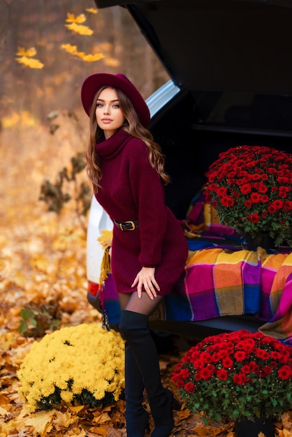 Beautiful girl in cosy knitted burgundy dress and hat sitting on nature with autumn background