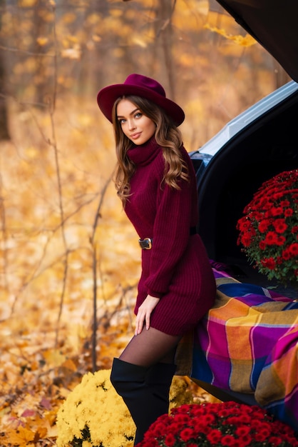 Beautiful girl in cosy knitted burgundy dress and hat sitting on nature with autumn background.