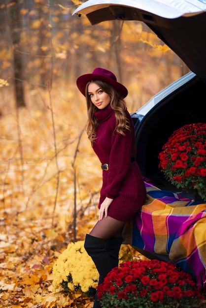 Beautiful girl in cosy knitted burgundy dress and hat sitting on nature with autumn background