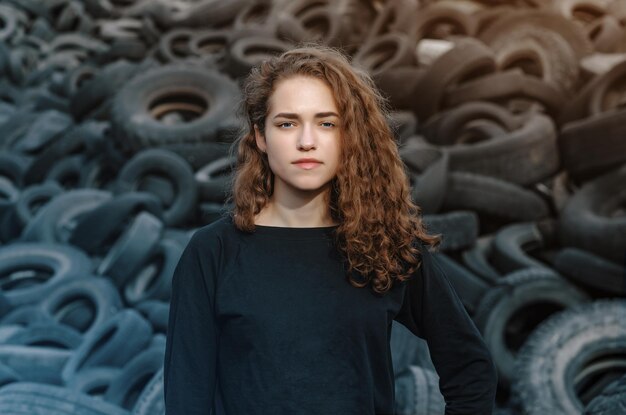 Photo beautiful girl curly hair portrait on the background of a dump of old car tires looking at camera