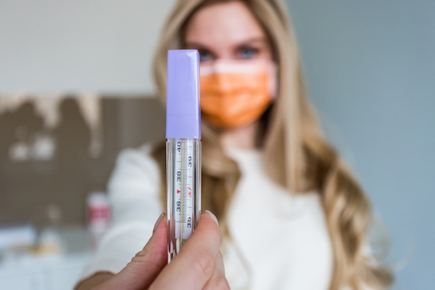 Foto bella ragazza dottore in una mascherina medica tiene in mano un termometro e lo mostra alla telecamera.