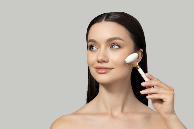 A beautiful girl does facial massage with a roller on a gray background