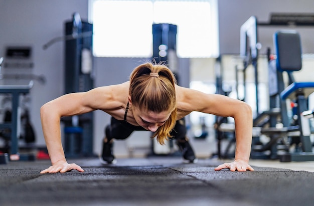 Beautiful girl does pushups from the floor for training the muscles of the hands in the gym on the background of sports equipment Sportswoman with hair in ponytail is doing muscle training