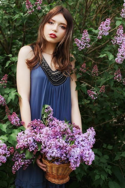 Bella ragazza in un vestito che propone vicino a un cespuglio di lillà in una giornata estiva, fiori viola nel parco. ritratto di primavera di una ragazza in natura al sole