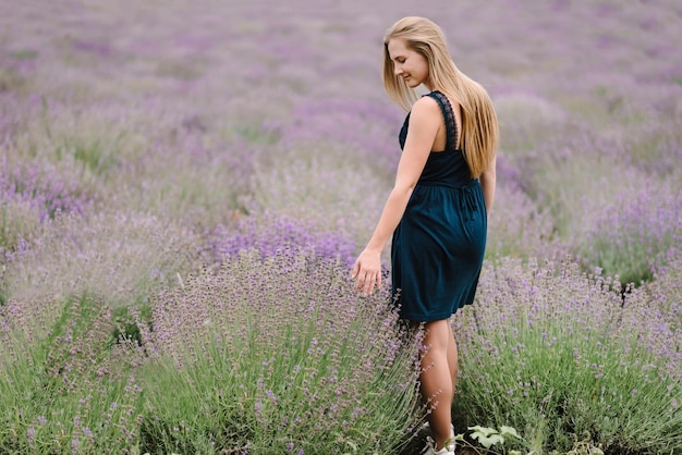 보라색 라벤더 밭에서 드레스를 입은 아름다운 소녀 라벤더 밭을 걷는 아름다운 여자 소녀는 라벤더를 수집합니다 꽃 숲 사이의 빈터 여름 자연을 즐기십시오 후면보기