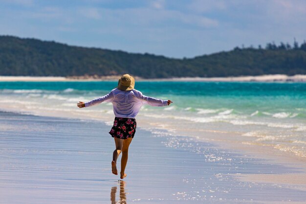 a beautiful girl in a dress, shirt and hat spreads her arms in joy walking along whitehaven beach