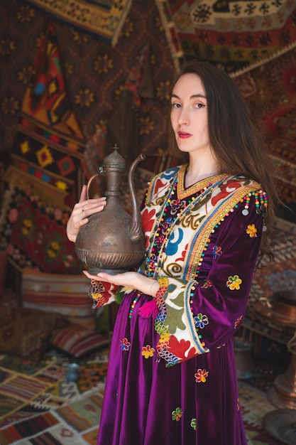 Beautiful girl dressed in Turkish national clothes in the room with the interior there are many carpets on the walls with a teapot in his hands