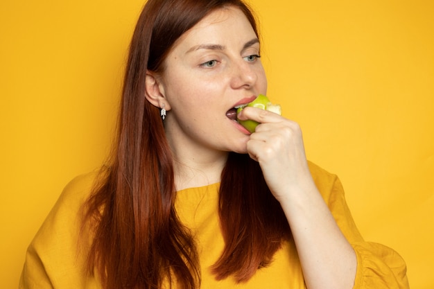 노란 벽 벽에 서있는 녹색 사과 먹는 아름 다운 소녀. 다이어트와 적절한 영양 개념.