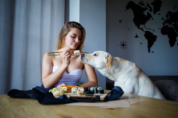 Photo beautiful girl eating sushi close-up with her dog labrador, smiling, holding a sushi roll with chopsticks. healthy japanese food.