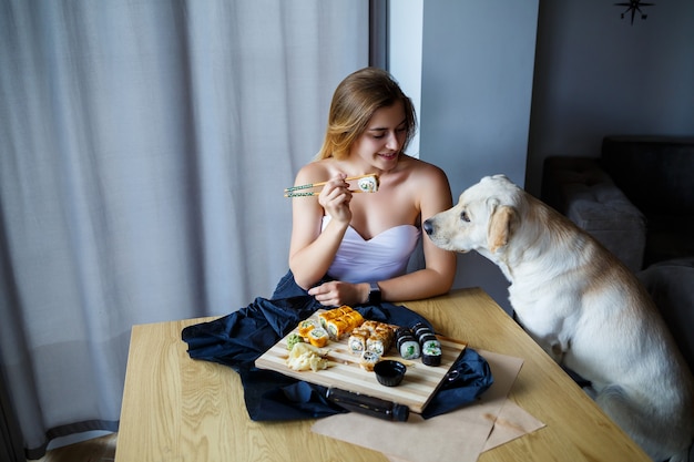 Beautiful girl eating sushi close-up with her dog labrador, smiling, holding a sushi roll with chopsticks. Healthy Japanese food.