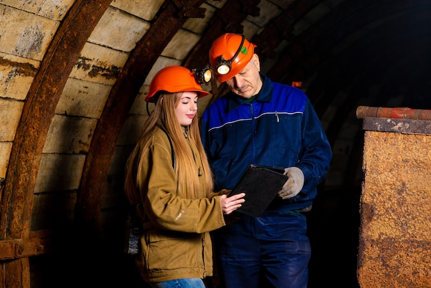 A beautiful girl and an elderly man in a protective suit and helmet are standing in a mine with a tablet in their hands