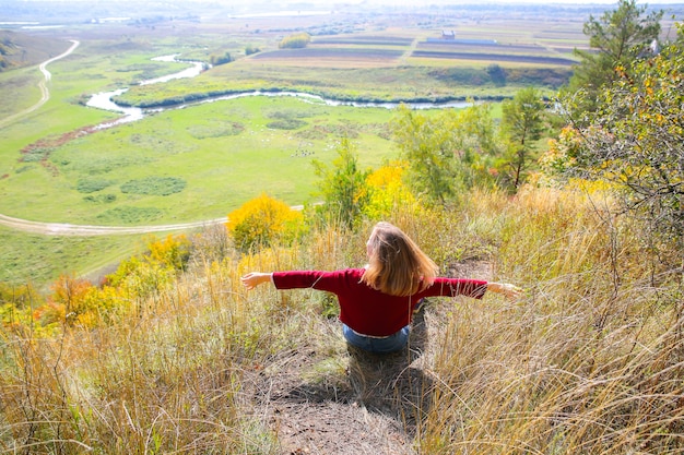 Beautiful girl enjoying nature view. Hipster on the hill. River and field below. Autumn season. Stylish woman outdoors. Tourist concept. Travel discover the world.