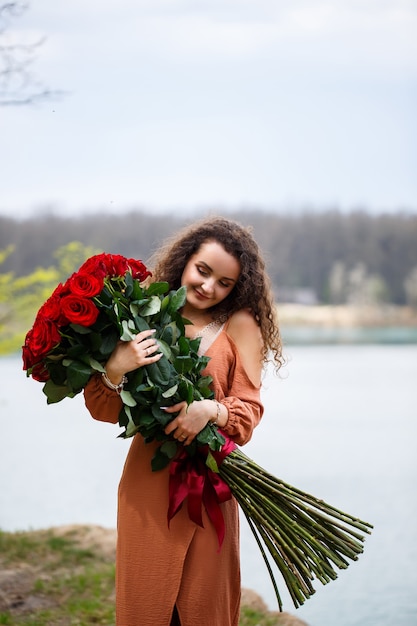 A beautiful girl of European appearance with curly hair and a smile on her face with a huge bouquet of red roses on a background of blue lake. Warm summer day, happy young woman, emotions of joy