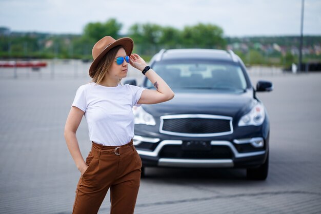 A beautiful girl of European appearance with glasses and a brown hat is standing near a black car.