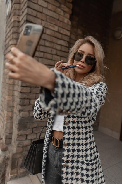 Beautiful girl in fashionable clothes smokes a vape cigarette and takes a selfie on a smartphone in the city