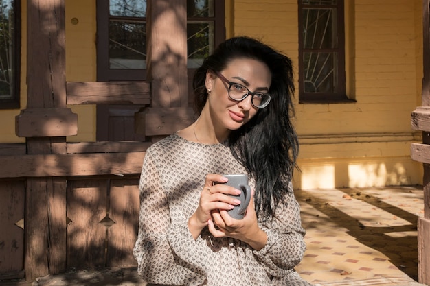 Una bella ragazza con gli occhiali sta riposando e bevendo il tè, seduta sulla veranda autunnale.