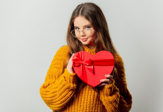 Beautiful girl in glasses and yellow sweater with heart shape gift box on white background