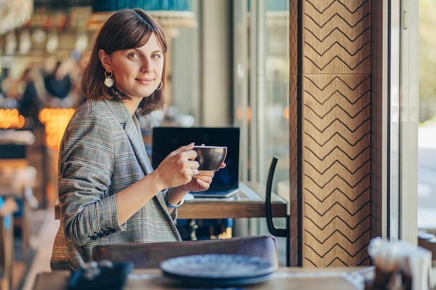 Beautiful girl in gray blazer, drinking coffee and working on net-book   in cafe. Freelancer working in coffee shop. Learning online.