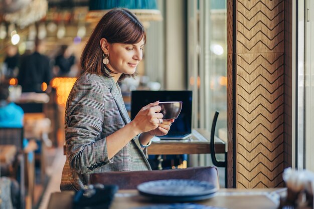 Beautiful girl in gray blazer, drinking coffee and working on net-book   in cafe. Freelancer working in coffee shop. Learning online.