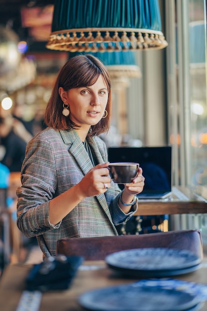 Bella ragazza in giacca grigia, bere caffè e lavorare su net-book nella caffetteria. libero professionista che lavora in una caffetteria. imparare in linea.