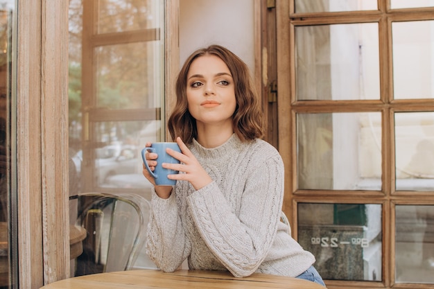 灰色のセーターを着た美しい少女がコーヒーショップで朝のコーヒーやココアを飲む秋の休息