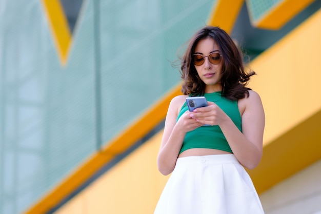 Beautiful girl in a green top and white shorts holds a mobile phone in her hands and looks into it Against the background of a yellow building