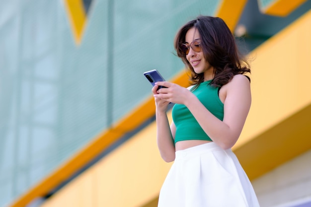 Beautiful girl in a green top and white shorts holds a mobile phone in her hands Smiling and looking at the phone Against the background of a yellow building