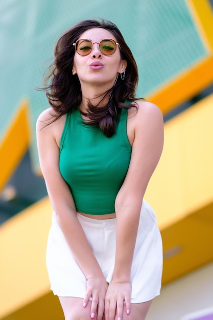 Beautiful girl in a green top and white shorts poses making a kiss to the camera Against the background of a yellow building