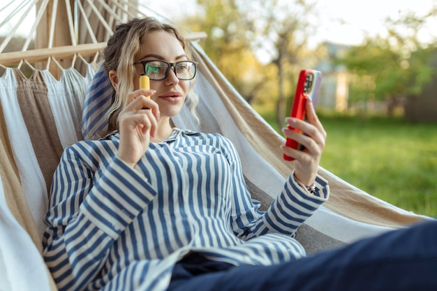 A beautiful girl in a hammock smokes an electrode cigarette and uses a smartphone outdoor