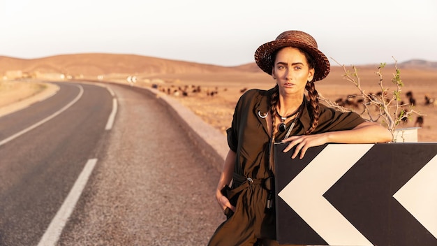 Beautiful girl in a hat near a road sign. In the background is a road in the desert .Morocco