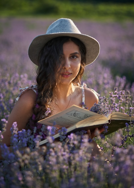 Beautiful girl in hat sitting in purple lavender field and read book.