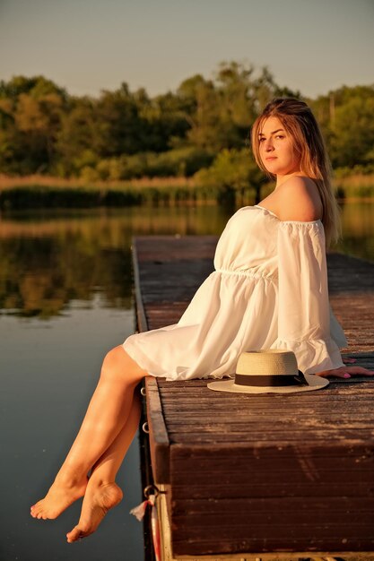 A beautiful girl in a hat stands on the shore hat in hand