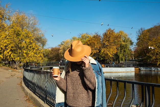 帽子をかぶった美しい女の子がコーヒーを飲んで秋の公園を歩いています