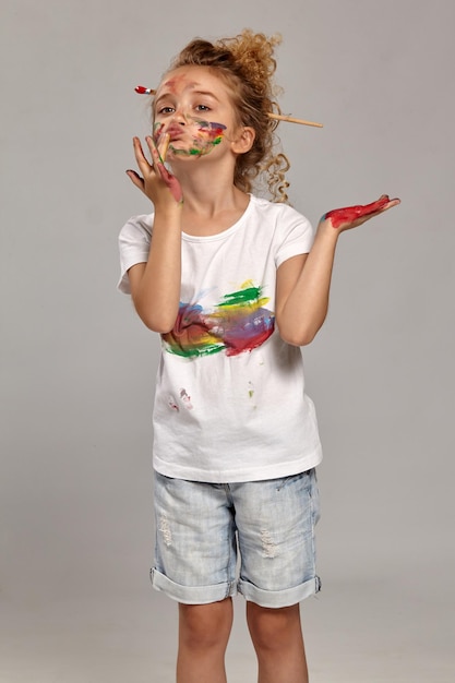 Beautiful girl having a brush in her lovely haircut, wearing in a white smeared t-shirt. She is posing with a painted hands and cheeks, pains her lips, on a gray background.