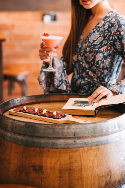 Bella ragazza che tiene uno squisito cocktail cremoso in un bicchiere speciale al bar si siede su un tavolo a forma di botte di legno