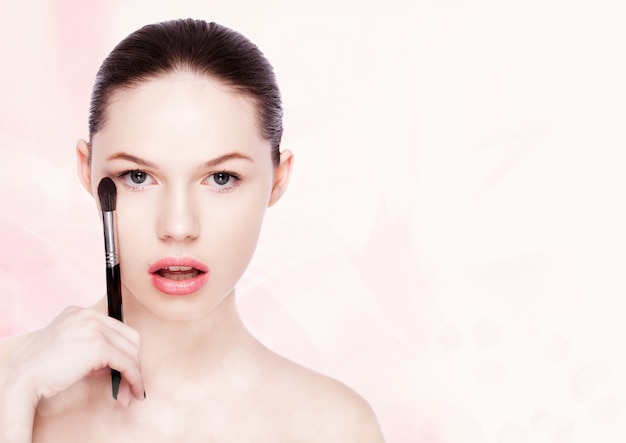 Beautiful girl holding makeup brush for foundation powder on pink bokeh background