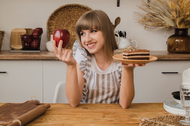 A beautiful girl holds a cake in one hand and an apple in the other and thinks what to eat