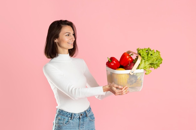 美しい少女は、ピンクの背景に健康野菜グラスファイバーのバスケットを差し出します