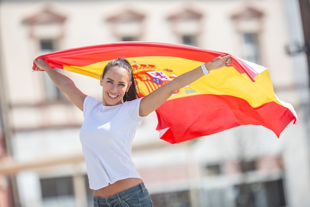 Foto la bella ragazza tiene una bandiera spagnola nelle sue mani dietro di lei, tifo per una strada.