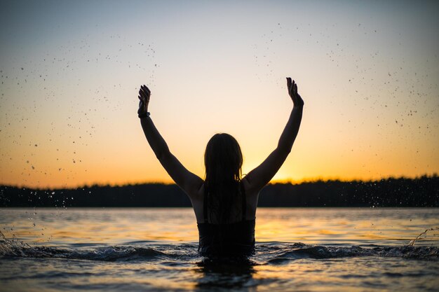 写真 長い黒い水着を着た美しい女の子が夕暮れや夜明けの光で湖で泳いでいます