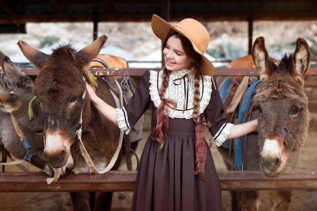 写真 レトロなドレスとロバに餌をやるロバ農場の帽子で美しい少女。収穫。秋の時間。