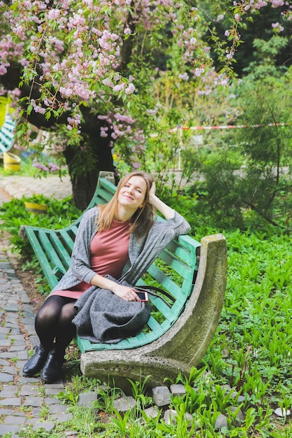 Красивая девушка сидит на скамейке в саду под цветущим деревом. Цветут сакуры на открытом воздухе. Весеннее настроение. Женщина в розовом платье. Расслабляющий.