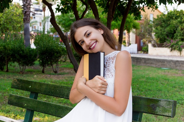 Beautiful girl is sitting on the bench in the park and hugging a book