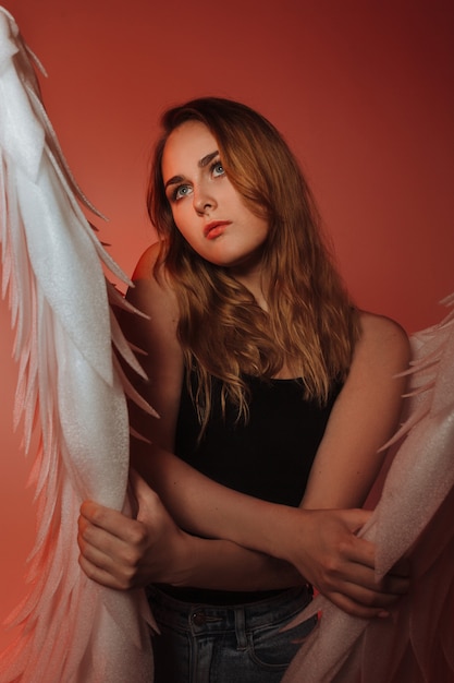 A beautiful girl is standing in jeans with big white wings on a red background