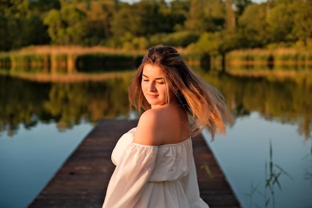 A beautiful girl is standing on the pier in the background there is a river a lake